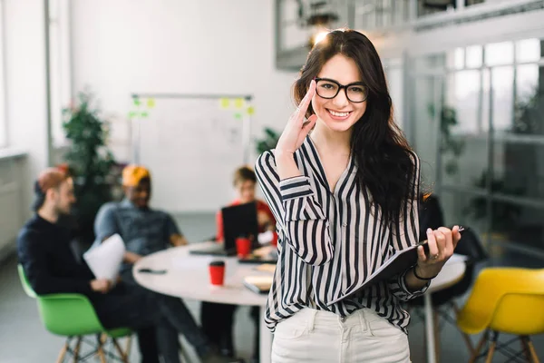 Belle femme d'affaires brune branchée dans des lunettes avec un dossier de documents dans ses mains à l'intérieur. Équipe de jeunes travailleurs discutant sur le fond — Photo