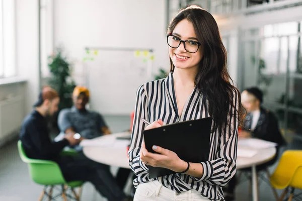 Young attractive designer woman in glasses smiling at camera in creative office while her colleagues sitting at the table and talking on the background — 스톡 사진