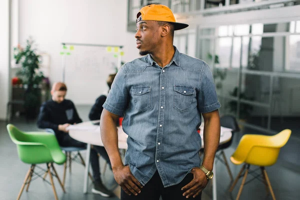 陽気なアフリカ系アメリカ人のヒップスターの男は、自由な時間の間にロフトオフィスに立って、幸せな暗い肌の男は、彼の仕事を楽しんでいます。オフィスで新しいプロジェクトで働くフリーランサーの若いチーム. — ストック写真