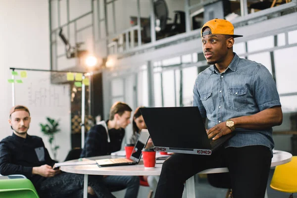Colega afro-americana de pele escura em camisa jeans e boné amarelo e usando laptop no escritório moderno. Multirracial jovens criativos estão trabalhando em conjunto com laptop, tablet, telefone inteligente . — Fotografia de Stock