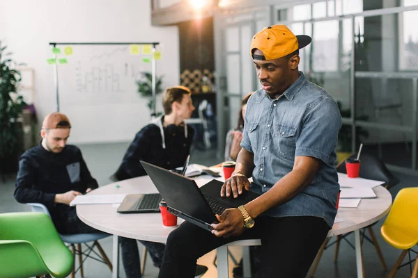 Colega afro-americana de pele escura em camisa jeans e boné amarelo e usando laptop no escritório moderno. Multirracial jovens criativos estão trabalhando em conjunto com laptop, tablet, telefone inteligente . — Fotografia de Stock