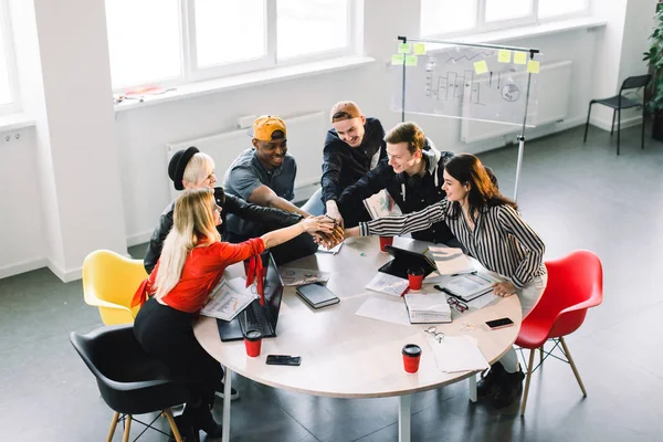 Team communication. Top view part of group of six young people in casual wear discussing something with smile while sitting at the office table and holding arms together