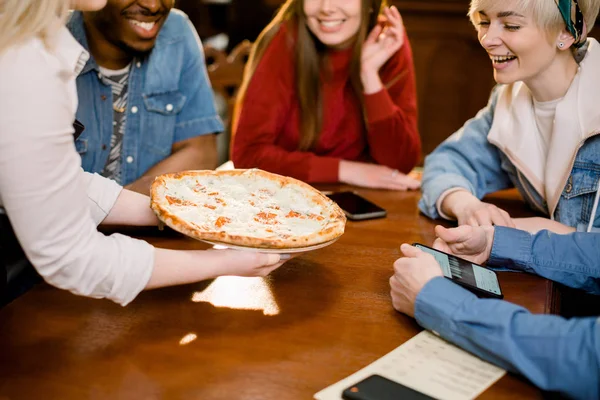 Young Caucasian woman waitress bringing pizza for group of diverse multiracial friends in cafe.