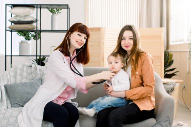 Genç, gülümseyen kadın doktor küçük kızı evde annesinin kollarında muayene ediyor.