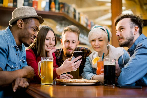 Kapalı bir kafede pizza paylaşan genç arkadaşlar. Multiracial mutlu bin yıllık arkadaşlar birlikte pizza yemek, komik şaka gülerek ve akıllı telefon kullanarak eğlenmek — Stok fotoğraf