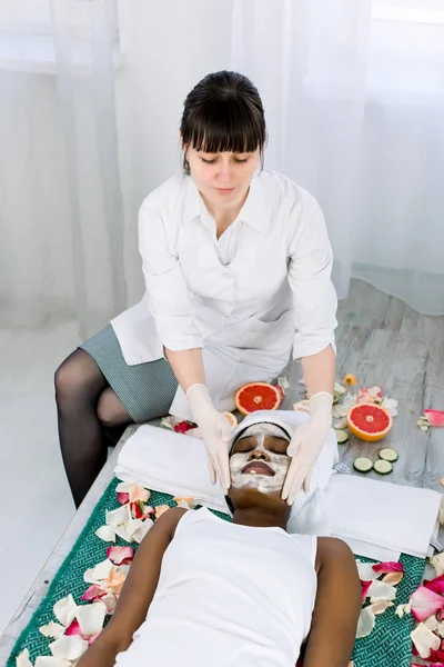 Face peeling mask, spa beauty treatment, skincare. Pretty African woman getting facial care by beautician at spa salon