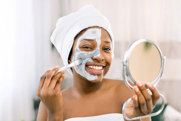 Wanita Afrika mengenakan handuk, dengan masker wajah. Close-up Of African Woman Applying Facial Mask In Spa — Stok Foto