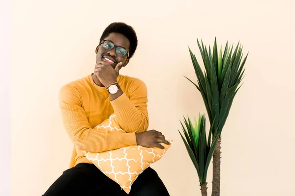 Inteligente y ambicioso. Joven hombre africano confiado mirando a la cámara y sosteniendo la mano en la barbilla mientras está sentado en la silla contra el fondo amarillo con planta tropical — Foto de Stock