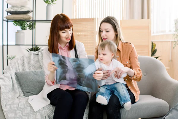 Mujer bonita médico de familia mostrando imagen de rayos X a madre joven con niña alegre en sus manos — Foto de Stock