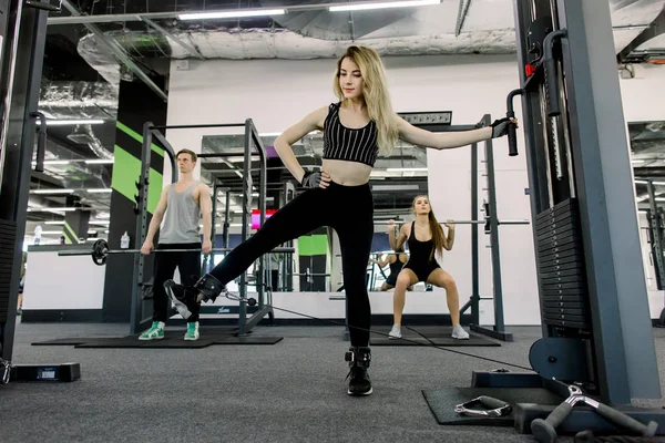 sport, bodybuilding, lifestyle and people concept - young girl doing leg stretching, young man and woman with barbell flexing muscles and making shoulder press squat in gym