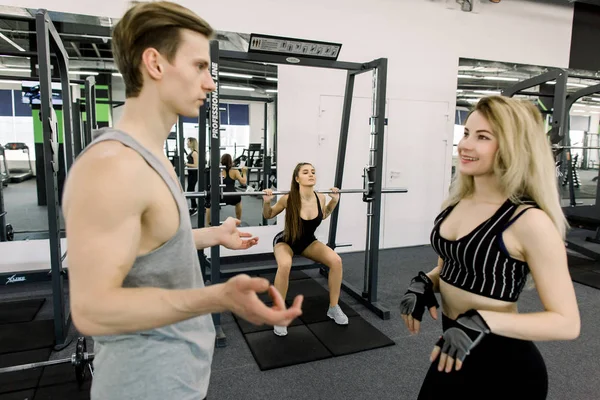 Fit jonge vrouw hijs barbells op zoek gericht, trainen in een sportschool met haar vrienden, juichen haar op en praten — Stockfoto