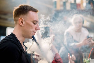 Siyah tişörtlü kafkas genç adam arkadaşlarıyla birlikte kafede masada otururken nargile içiyor