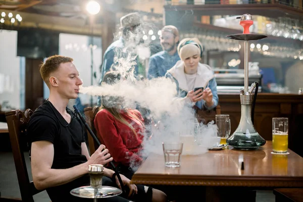 Amigos multiétnicos de grande grupo meninos e meninas sentados à mesa no café, bebendo cerveja e fumando narguilé . — Fotografia de Stock