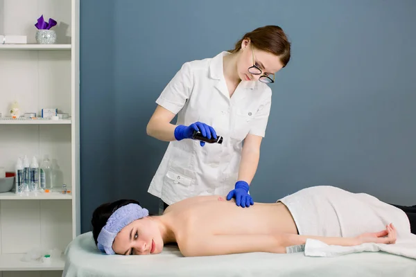 Female beautician doctor with woman patient in spa wellness center. Professional massagist make body back oil massage to beautiful Caucasian girl in cosmetology cabinet or beauty parlor.