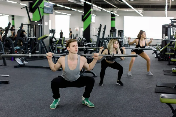 Jóvenes haciendo ejercicio con campanas en el gimnasio. Las mujeres atractivas y el entrenador masculino musculoso guapo están entrenando en el gimnasio moderno ligero. Hermosa chica sentadillas bajo la supervisión del entrenador . —  Fotos de Stock