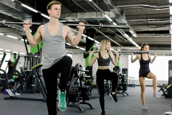 Deporte, culturismo, estilo de vida y el concepto de la gente - hombre joven y dos mujeres con los músculos de flexión de la barra y hacer que la prensa de hombro en cuclillas en el gimnasio —  Fotos de Stock