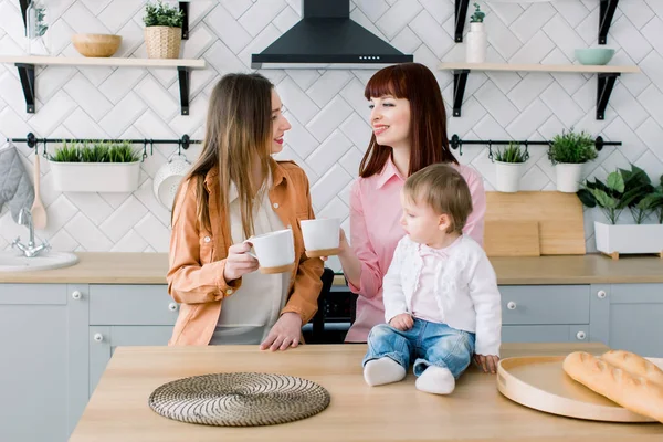 Két vidám fiatal nő ül egy asztalnál csészéket a konyhában. Kis kislány ül az asztalon. Reggeli, barátság koncepció — Stock Fotó