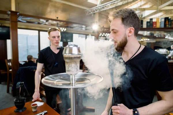 Dois garotos preparando a shisha colocando os carvões no topo. Um homem acendendo narguilé no fundo bar . — Fotografia de Stock