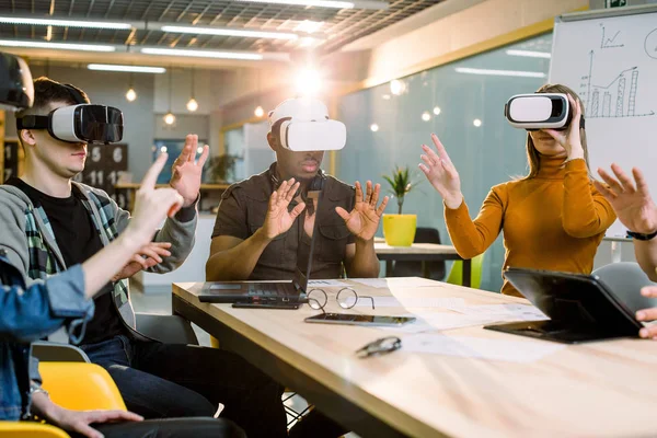 Young men and women sitting at a table with virtual reality goggles. Multiethnical business team testing virtual reality headset in office meeting.