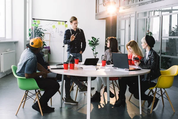 Jeunes créatifs multiraciaux dans le bureau moderne travaillent ensemble avec ordinateur portable, tablette, téléphone intelligent, ordinateur portable. Équipe hipster réussie dans le coworking. Jeune homme près du mur de verre ayant la parole — Photo