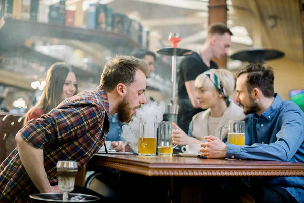Şık sakallı yetişkin bir adamın bir kafenin kapalı mekanında nargile içerken, arkadaşlarıyla birlikte masada otururken çekilmiş yakın çekim. — Stok fotoğraf