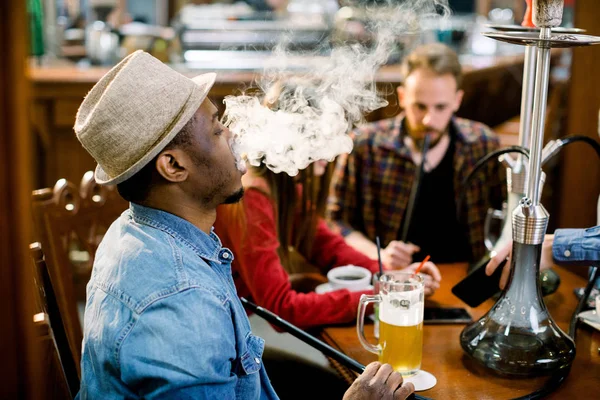Cheerful company of friends, drinking beer and smoking hookah in shisha lounge. Rest in hookah, African man makes tornado out of smoke shisha