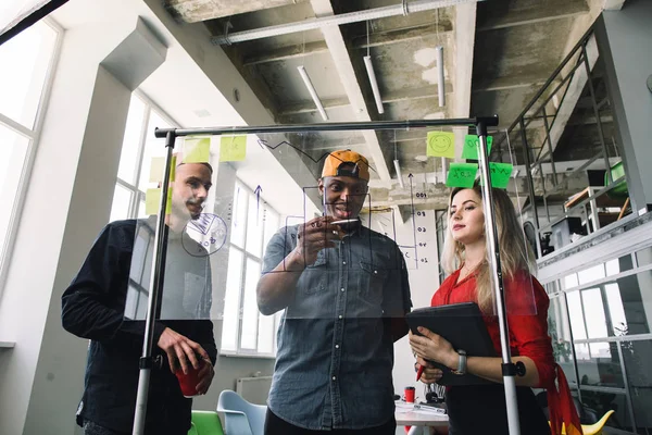 Trois hommes d'affaires multiethniques en tenue décontractée discutant et planifiant le concept, travaillant ensemble dans le bureau loft avec mur de verre. Face avant du marqueur mural en verre et des autocollants. Bureau de démarrage . — Photo