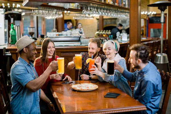 Happy five friends having drinks in a bar, They are sitting at a wooden table with beers and pizza.