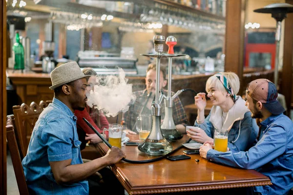 Cheerful company of friends, drinking beer and smoking hookah in shisha lounge. Rest in hookah, African man makes tornado out of smoke shisha