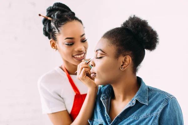 Jovem bela mulher afro-americana aplicando maquiagem profissional por artista de maquiagem africana. Estúdio — Fotografia de Stock