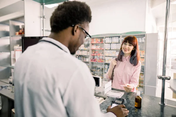 Mladá Kavkazská žena, která platí pro medicieny kreditní kartou v lékárně. Farmaceutika pro mladé ženy v lékárně u Afričana — Stock fotografie