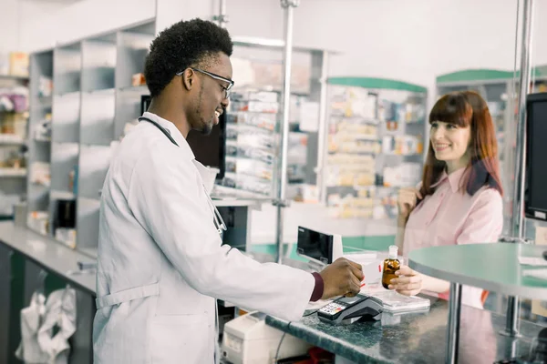 Mladá Kavkazská žena v lékárně platí za medicieny kreditní kartou. Farmaceutika pro mladé ženy v lékárně u Afričana — Stock fotografie