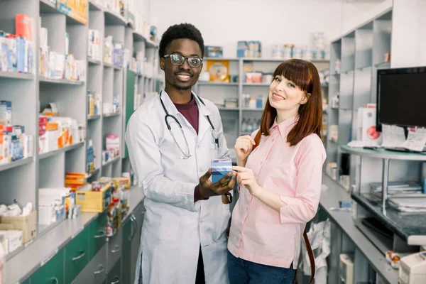 Heureuse jeune cliente debout près d'un pharmacien homme africain, tenant la médecine ensemble. Belle femme regardant la caméra et souriant. Africain en manteau blanc aider les clients en pharmacie . — Photo