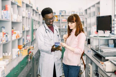 Eczanede önerilen ilaçların memnun genç kız müşteri, modern eczanede Afrikalı adam eczacı ile birlikte ayakta iken