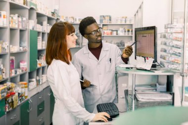 Eczanede iki genç eczacı ekibi ilaçlar ile rafların önünde duran ve ilaçlar bilgisayar yapma emri üzerinde çalışıyor