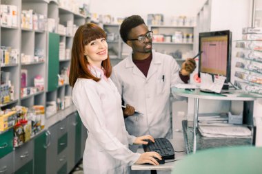 Modern eczane iç bir gülümseyen kadın eczacı ayakta portre, arka planda bilgisayar ile çalışan erkek meslektaşım.