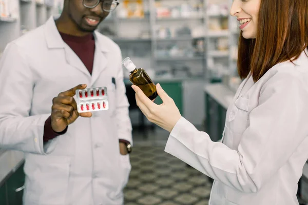 Closeup vágott portré két mosolygó barátságos gyógyszerészek dolgozó modern gyógyszetár. Afrikai ember tart vörös tablettát buborékfólia és kaukázusi nő rendelkezik üveg szirup — Stock Fotó