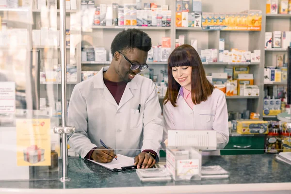 Farmacéuticos masculinos y femeninos multiétnicos confiados en farmacia. Hombre afroamericano farmacéutico tomando notas en portapapeles durante el inventario en farmacia — Foto de Stock