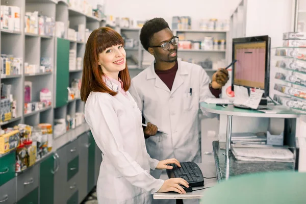 Portret uśmiechniętej kobiecej farmaceuty stojącej we wnętrzu nowoczesnej apteki, męski kolega pracujący z komputerem w tle. — Zdjęcie stockowe