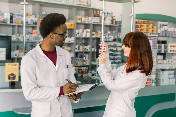 Africký muž zápis Farmakist do schránky zatímco jeho kolega má v rukou puchýř červených prášků, kontrola kvality léků — Stock fotografie