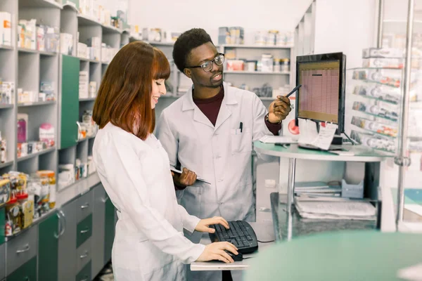 Zespół dwóch młodych aptekarza w aptece stojącej przed półkami z narkotykami i pracujących na porządku komputerowym leków — Zdjęcie stockowe