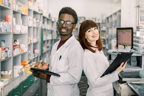 African man and Caucasian woman pharmacists are posing near table with cashbow in apothecary.