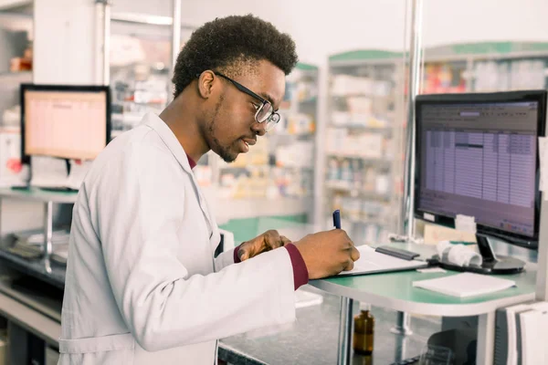 Farmacéutico afroamericano experimentado trabajando en PC en farmacia moderna y escribiendo notas de trabajo —  Fotos de Stock