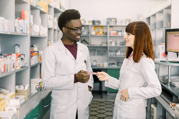 Pewnie mężczyzna i kobieta farmaceuci w aptece. African mężczyzna i kaukaski kobieta trzyma blister tabletek stojąc w nowoczesnej aptece — Zdjęcie stockowe