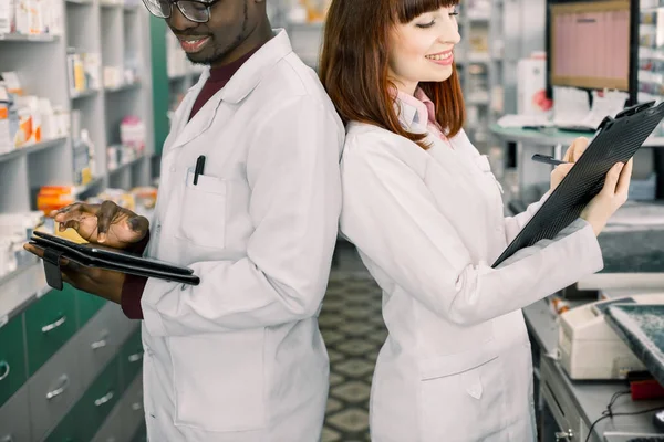 약국에서 일하는 두 명의 다인종 약사가 뒤로 서 있습니다. 약국에서 의약품 재고를 확인하는 남성 및 여성 약사. 클립 보드에 태블릿과 여자 쓰기를 사용하는 남자 — 스톡 사진