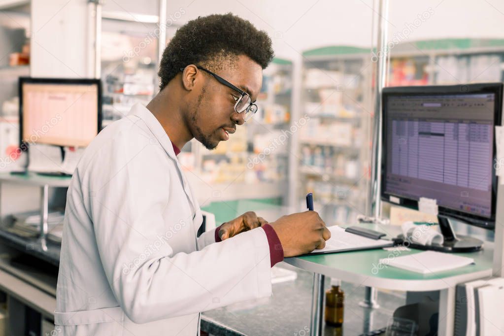 Experienced African American male pharmacist working on pc in modern pharmacy and writing working notes