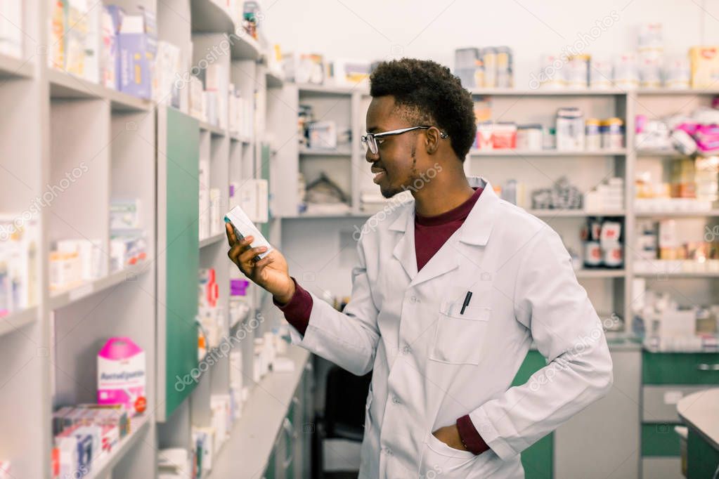 Portrait of African American male pharmacist standing in interior of pharmacy and looking at the medicine in hand