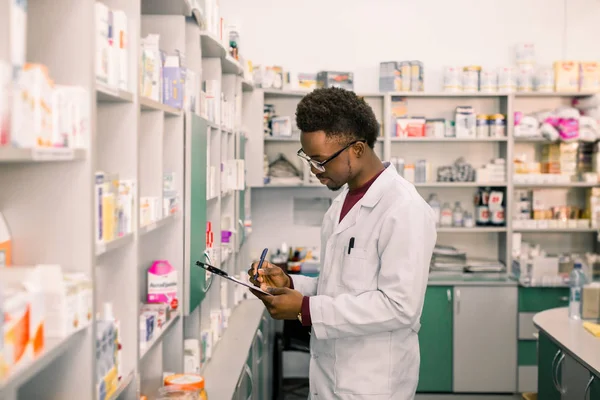 Młody African American mężczyzna farmaceuta stojący we wnętrzu apteki. Człowiek specjalista apteki dokonywania notatek w schowku podczas inwentaryzacji — Zdjęcie stockowe