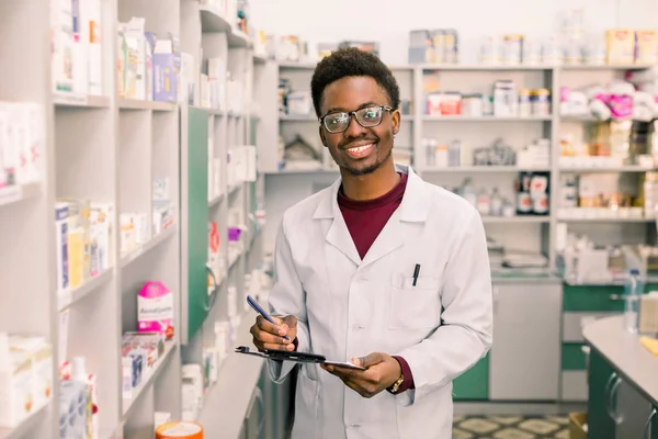 미소 아프리카계 미국인 남자 약사 또는 화학자 쓰기 에 클립 보드 동안 서 있는 동안 의 내부 의 약국 — 스톡 사진
