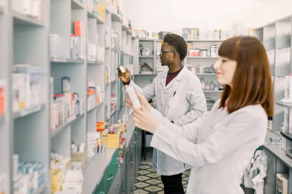일하는 두 약사. 백인 여자와 아프리카 남자 착용 특별 한 의료 유니폼. 알 약으로 패키지를 보여주는 여자. 배경에 약을 찾는 남자. — 스톡 사진